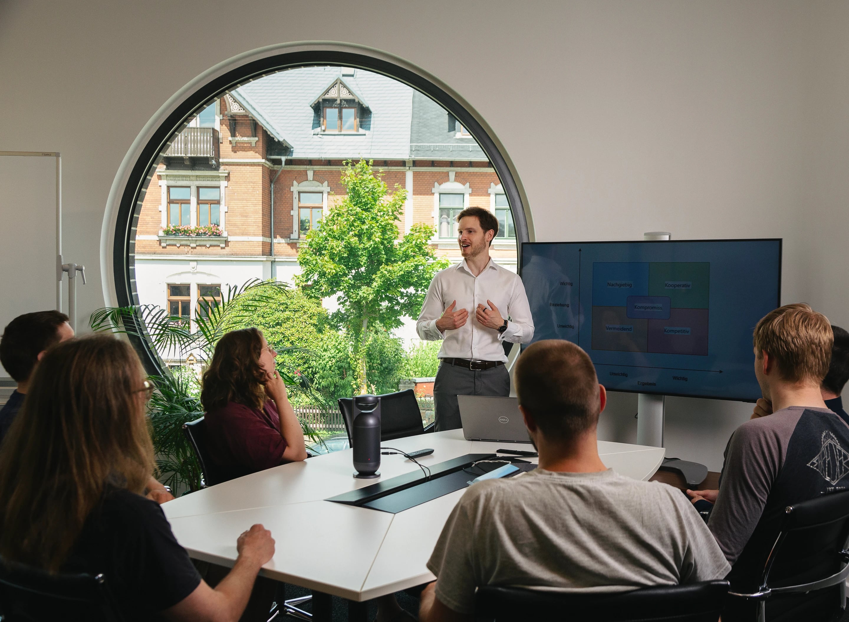 Seminarraum mit rundem Fenster; der Seminarleiter spricht vor der Gruppe am Konferenztisch.
